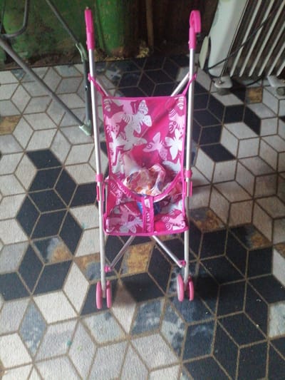 A pink doll stroller with a butterfly pattern is centrally positioned on a patterned floor. The stroller is made of metal with pink accents and wheels. It appears to be empty except for some crumpled fabric inside the seat. The background is blurred, but hints of a green surface and a white ribbed object are visible. The floor has a geometric pattern of black, white, and grey cubes. The image appears to be taken indoors. The location is likely the city of Степной, Russia.