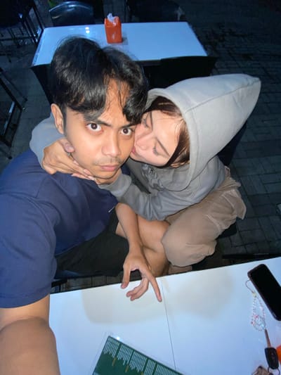A high-angle, close-up, outdoor photograph captures a young couple posing for a selfie. The man, wearing a navy blue long-sleeved shirt and dark shorts, is positioned in the foreground, looking directly at the camera with wide eyes and a slightly questioning expression. A woman, wearing a grey hooded sweatshirt and tan pants, is leaning in from his right side, kissing him on the cheek. Her arms are wrapped around his neck and shoulders. They are seated at a white table, with another white table visible in the background behind them, where an orange tissue box sits. The ground beneath them appears to be paved with interlocking bricks. The lighting suggests it is evening or nighttime, with artificial lights illuminating the scene. On the white table in the foreground, a smartphone, a keychain with colorful beads, and a car key are visible. In the lower right corner, a green and white menu is partially visible. The location is identified as Mekar Bakti, Indonesia.