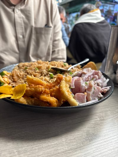 An angled, close-up shot captures a vibrant Peruvian meal served on a large black platter, likely in a restaurant setting in Jesús María, Peru. The dish is a "combinado" featuring three distinct components.

On the upper left side of the platter, there is a generous portion of ceviche, characterized by tender chunks of light-colored fish, thinly sliced red onions, and a delicate sprinkle of green herbs, possibly cilantro. Adjacent to the ceviche, a substantial serving of arroz chaufa, a Peruvian fried rice, is visible. The rice appears fluffy and golden-brown, mixed with small green pieces like peas or scallions. A silver spoon is partially submerged in the rice.

To the right, a pile of golden-brown fried seafood, likely chicharron de calamar (fried calamari) or fish, is arranged. These pieces have a crispy texture. Bright yellow, thin plantain chips garnish the dish, adding a pop of color and an additional texture.

The table surface beneath the platter is a light-colored wood grain. In the background, blurred figures suggest other people in the restaurant. One individual, seen from behind and mostly out of focus, wears a black jacket with a white and yellow collar. Another person, likely the diner taking the photo, is partially visible in the bottom left, wearing a light tan or beige collared shirt. The lighting indicates an indoor setting, likely during the day or under bright artificial lights. No visible text is present in the image.