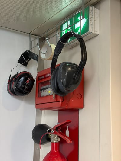 The image shows a close-up of safety equipment mounted on a white wall. From left to right, there are a pair of black and red earmuffs hanging from a metal rack. To the right of the earmuffs is a red electrical box with a transparent cover, showing several circuit breakers. Above the electrical box, a roll of tape is partially visible. Further to the right, another pair of black earmuffs is hanging. At the top of the image, a green emergency exit sign with a white arrow pointing right is illuminated. Below the emergency sign, a section of a red fire extinguisher is visible, with its handle and nozzle. The scene appears to be indoors, possibly in a utility area or workshop. The location is not specified as Arles, France; the image content does not provide any clues to this location.