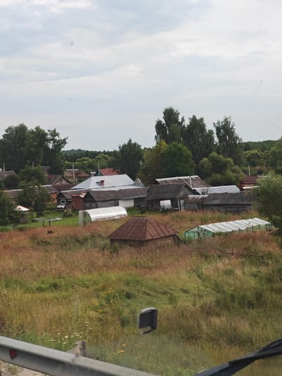 The image depicts a rural village scene in what is identified as Кувак-Никольское, Russia. The landscape features a collection of houses with various roofing materials, including metal and dark shingles. Some houses appear older and weathered, constructed from wood and brick. There are also greenhouse structures visible in the foreground, suggesting agricultural activity. The surrounding environment is characterized by overgrown fields with dry grass and some green vegetation, indicative of a summer or early autumn setting. Trees are present in the background, forming a dense forest line. The sky is overcast with clouds. A metal railing is visible in the lower portion of the image, suggesting the photograph was taken from a moving vehicle or a raised vantage point. There are no people or animals clearly identifiable in the scene. The overall impression is one of a quiet, possibly remote, rural settlement.
