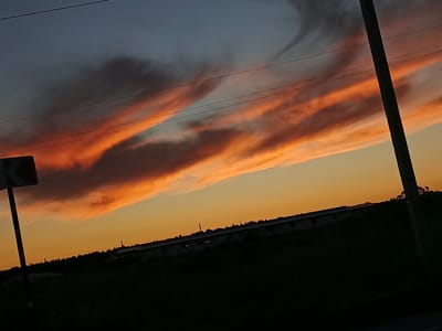 A photograph captures a dramatic sunset over a rural landscape, likely on the outskirts of Воскресенское, Russia. The sky is a gradient of deep blue at the top, transitioning to a vibrant orange and yellow near the horizon. Wispy clouds, illuminated by the setting sun, streak across the sky in fiery shades of orange and pink. Three parallel power lines stretch horizontally across the upper portion of the image.
In the foreground, a dark silhouette dominates the lower half of the frame. On the left, a signpost with a white chevron on a dark background is partially visible. A dark, vertical pole, likely a utility pole, stands prominently on the right side of the image. The horizon is marked by a dark, undulating line of trees and low structures, possibly agricultural buildings or a small settlement, silhouetted against the bright sky. The ground in the immediate foreground is also a dark silhouette, suggesting dense vegetation or a shadow. The overall impression is one of a serene yet striking natural phenomenon.