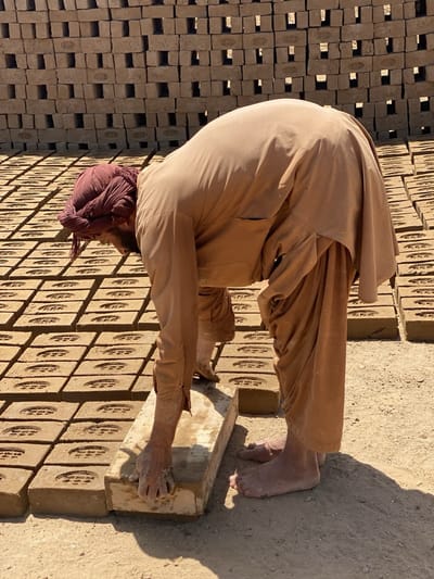 In Deh-E Bagh, Afghanistan, a man wearing a tan tunic and maroon turban is working in a brickyard. He is barefoot and bending over, placing a long, light-colored brick onto a grid of similar, darker, brick-shaped objects. Behind him, stacks of finished bricks are visible. The ground is dry and dusty. The scene is brightly lit, suggesting a sunny day.