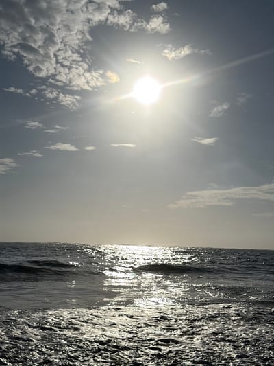 The image depicts a seascape, likely taken in Cabarete, Dominican Republic. The right side shows the ocean, with dark, choppy waves reflecting sunlight. The left side shows a sky with clouds and a bright sun, contributing to the glare reflected on the water.