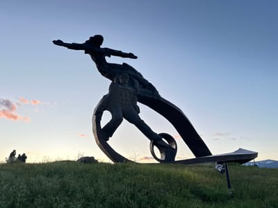 The image depicts a large, dark-colored sculpture of a man and a woman on a grassy hill under a blue sky. The woman is positioned above the man, both with outstretched arms. The sculpture incorporates circular and curved shapes. To the left, in the distance, are several silhouetted figures standing on the hillside. In the background, there are mountains and a light fixture positioned under the sculpture. This is located in the city of Unknown, Armenia.