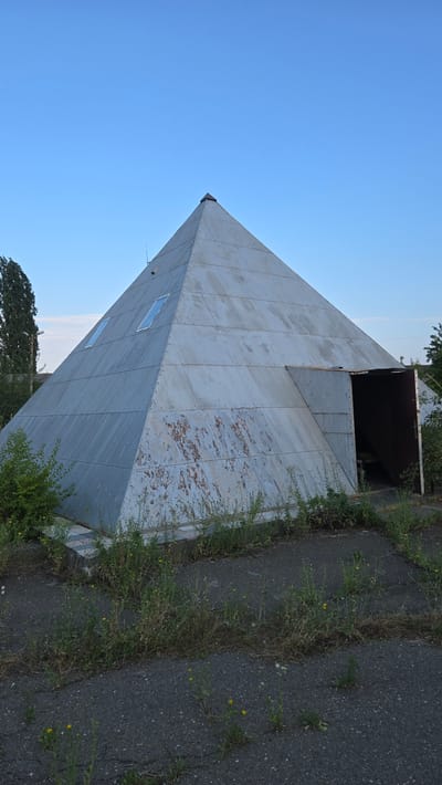 A metallic, pyramid-shaped structure stands in an overgrown, unpaved area under a clear blue sky. The structure is clad in overlapping metal panels, some of which show signs of rust and wear. Two rectangular windows are visible on one of the faces. An open doorway on the right side reveals a dark interior with steps leading up. Greenery, including tall grass and bushes, surrounds the base of the pyramid and encroaches onto the cracked asphalt ground in the foreground. A tall, slender tree is visible to the left of the structure, and other trees and vegetation can be seen in the background. The scene appears to be located in a relatively isolated or abandoned area, possibly on the outskirts of Pervomaisk, Moldova.