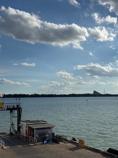 The image depicts a waterfront scene with a ferry dock in the foreground and a body of water stretching to the horizon under a partly cloudy sky. On the dock, a small white building with a corrugated metal roof stands near a heavy-duty crane. Several people are visible on the dock, including one individual standing near a collection of blue and green plastic containers. A yellow sign with Thai text and the word "PHANGAN" is positioned on the edge of the dock. The water is a calm, greenish-blue, with small waves. In the distance, a tree-lined shore is visible, with a distinctive rocky outcrop and a communication tower silhouetted against the sky. Two small boats are present on the water, further enhancing the sense of a coastal or riverine environment. The overall atmosphere suggests a location within the Tambon Don Sak district of Thailand, likely a ferry terminal.