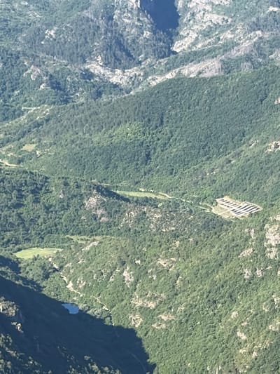 The image is an aerial view of a mountainous, forested region. The terrain is characterized by steep slopes covered in dense green vegetation, with visible rocky outcrops. In the mid-ground, a cluster of what appear to be solar panels is situated on a cleared area of the hillside. Further to the left and lower in the frame, a small body of water, possibly a lake or pond, is visible, reflecting the dark shadow of the mountain. The overall impression is one of a natural landscape with some evidence of human intervention. The location is specified as Kućišta, Montenegro.