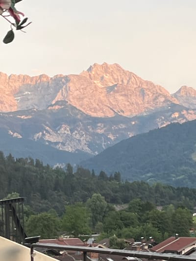 A photograph captures a scenic mountain vista with a foreground showing rooftops in Garmisch-Partenkirchen, Germany. The mountains are bathed in the soft, warm light of either sunrise or sunset, highlighting their rugged textures and snowy peaks. In the midground, lush green forests cover rolling hills. The foreground features a few buildings with red-tiled roofs, characteristic of the Bavarian architectural style. A dark metal railing is also visible in the lower left corner, suggesting the photo was taken from a balcony or terrace. No people are visible in the frame.