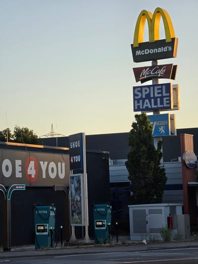 The image depicts an outdoor scene in Bischofsheim, Germany, featuring a collection of businesses and infrastructure. Prominently displayed is a tall McDonald's sign with the iconic golden arches and branding for McDonald's and McCafé. Below these, signs for "SPIEL HALLE" (Game Hall) and "HESSEN" with a lion emblem are visible. To the left, a large sign reads "OE 4 YOU," suggesting a retail store. In the foreground, there are several Electra charging stations, indicating an area with electric vehicle charging facilities. The background includes a large building, possibly a commercial complex, with a dark facade and metallic accents, and a glimpse of a power transmission tower in the distance. The environment appears to be an urban or commercial area, possibly an industrial park or a shopping district, with a road in the immediate foreground. The sky is clear and shows the soft light of late afternoon or early morning. There are no people visible in the image.