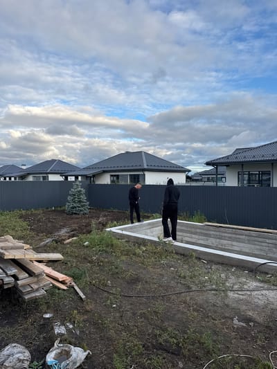 The image depicts a construction site in what is described as the city of Primorsky, Russia. In the foreground, a concrete foundation for a structure is visible, with two individuals standing near it. One person, wearing black clothing and a hooded sweatshirt, is standing on the edge of the foundation. The other person, also in dark clothing, is facing away from the camera and appears to be looking at something on the ground.
Behind the foundation, a dark gray metal fence runs horizontally across the frame, separating the construction area from a row of newly built houses. These houses have dark gray roofs and white walls, with modern architectural designs. A small, young evergreen tree is planted in the soil behind the fence, in front of the houses.
To the left of the foundation, a pile of wooden planks and construction materials is stacked on the ground. The ground itself is covered in dirt and grass, with some scattered debris, including a white plastic bag with water inside. The sky above is partly cloudy, with dramatic clouds suggesting potentially inclement weather. The overall scene conveys a sense of ongoing construction and development in a suburban or semi-rural setting.