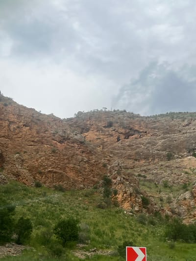The image displays a rugged mountainous landscape, attributed to the city of Arpi, Armenia, under an overcast sky. The upper portion of the frame is dominated by a light grey, cloudy sky, with some darker cloud formations visible towards the upper right. Below the sky, a large, stratified rock face forms the central subject, characterized by shades of reddish-brown, orange, and grey, indicating a weathered and possibly sedimentary geological composition. Sparse green vegetation, consisting of small bushes and shrubs, dots the rock face, particularly along its upper edges and in crevices. The lower third of the image features a vibrant green, grassy slope covered in various plants and small trees, suggesting fertile ground at the base of the mountains. In the bottom right corner, a portion of a red and white road sign is visible, displaying a white chevron pointing right, indicating a directional warning, likely for a curve in a road that is not visible. There are no people, animals, or other explicit man-made structures present in the scene.