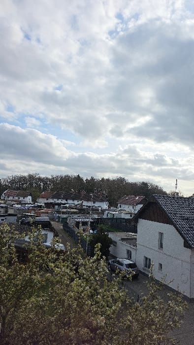 An overhead view captures a residential area in Heusden, Belgium, under a largely overcast sky with visible patches of blue. The foreground is dominated by the upper canopy of deciduous trees with light-colored blossoms, obscuring the ground level. In the midground, numerous residential structures are visible, primarily white-walled houses with dark-tiled, pitched roofs or flat roofs. A prominent white house with a dark tiled roof and visible windows is positioned on the right. Adjacent to this house, a light-colored sedan is parked in a driveway. Multiple unpaved or narrow roads/paths traverse the scene between properties. Streetlights and various fences are discernible. The background features a dense line of bare or sparsely foliated deciduous trees. No individuals or active interactions are observed.