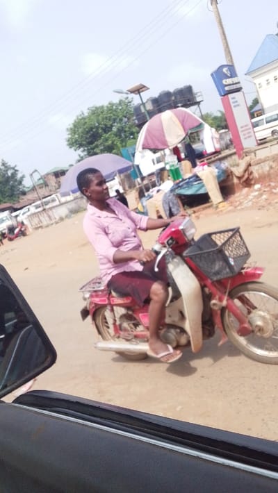 A forensic description of the scene in Igbo Ukwu, Nigeria, depicts an adult female operating a red underbone-style motorcycle on a light brown, possibly unpaved, road. The woman, with short dark hair, wears a light pink collared shirt, a dark red skirt, and white flip-flops. Her left foot is visible on the motorcycle. The motorcycle is equipped with a black wire basket on the front handlebars. The image is captured from inside a vehicle, with a dark window frame partially visible in the lower left foreground. In the mid-ground and background, a street market environment unfolds, featuring multiple vendor stalls shaded by striped umbrellas (one cream and maroon, another light purple). Merchandise, including clothing and bottled items, is visible at these stalls. A utility pole with power lines and a solar-powered street light stands alongside a blue and white commercial sign, possibly for "CRYSTAL MONEY PLUS," positioned near a building with a blue roof. Additional vehicles and structures are present further down the road under an overcast sky.