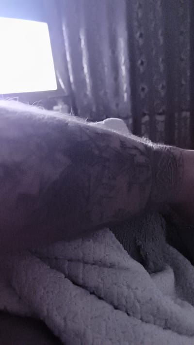 The image depicts a close-up, low-light view of a person's forearm with tattoos, resting on a textured, possibly plush, surface. A bright, rectangular light source, possibly a laptop screen or a light panel, is visible in the upper left corner, casting a glow on the scene. The background is dark and out of focus, showing vertical lines that could be curtains or blinds. The overall impression is one of a private, indoor setting, likely in a home. The location in Байқоныс ауылдық округі, Kazakhstan, is not visually identifiable from the image itself; this information is external to the visual content.
