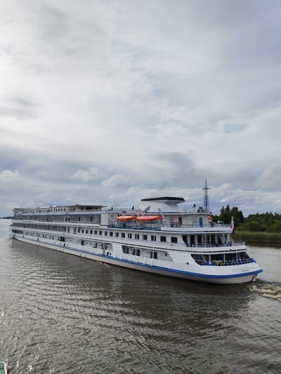 The image shows a large white cruise ship, with multiple decks, moving on a body of water. The ship has a blue stripe along its hull and a white superstructure with many windows. There are orange life rafts visible on one of the decks. The ship's name appears to be inscribed on the bow in Cyrillic letters. The water is choppy, with a wake trailing behind the ship. On the right side of the image, there is a shoreline with green trees and reeds. In the background, there is a tall pylon or transmission tower. The sky is overcast with scattered clouds. The scene is set on the river in the city of Kargulino, Russia.