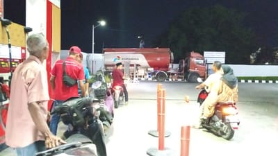 At a Pertamina gas station in Cotbadatunong, Indonesia, two men are seen at a gas pump, with one man in a pink shirt looking away from the camera and the other in a red shirt facing the camera, wearing a black fanny pack. A person in a red shirt and a peci hat is seen on a motorcycle, looking towards a large red Pertamina tanker truck. A man stands on top of the tanker truck, and another person is standing near the truck. Further in the background, a man and a woman ride a scooter. The scene is illuminated by artificial lights, and trees are visible in the dark background.