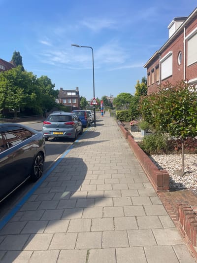 This is a photograph taken on a sunny day in Venlo, Netherlands. The image depicts a residential street scene with a focus on the sidewalk and parked cars. The perspective is from a low angle, looking down the sidewalk.
The subjects include several parked cars, a streetlamp, a brick building on the left, and trees and more residential buildings in the background. The sidewalk is paved with light-colored rectangular stones. A blue painted line runs along the curb, indicating a parking zone. The shadow of the streetlamp stretches across the sidewalk, indicating it is likely daytime.
Notable details include the clear blue sky with some wispy clouds, suggesting fair weather. The architecture of the brick building on the left, with its white-framed windows and roller shutters, is typical of Dutch residential areas. The trees are lush and green, indicating it is likely spring or summer. The license plate of the grey car in the foreground reads "ST-198-S".
There is no explicit activity or event taking place beyond the static scene of a quiet street. The overall mood is peaceful and ordinary, capturing a slice of everyday life in a suburban setting. The time of day appears to be late morning or early afternoon, given the length and angle of the shadows.
