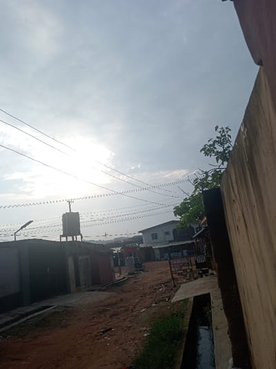 A hazy sky hangs over a street scene. Overhead power lines stretch across the view. A water tower rises above rooftops. A building is partially visible on the right, with some greenery near its top. A narrow drainage ditch runs alongside the road. The street itself appears to be dirt, with structures and shops on either side. A building dominates the right foreground, partially obstructing the view.