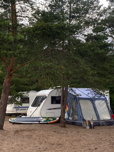 This is a media file featuring a caravan and an awning set up in a campsite, surrounded by pine trees. A paddleboard with a floral design and striped pattern stands upright next to the caravan. A camping stove and a small axe are visible on a log near the awning. The ground appears to be sandy. The scene is likely set in Yermilovo, Russia, given the pine forest and camping setup. The lighting suggests it might be daytime, possibly overcast, as the sky is bright but not intensely sunny. There is no visible text in the image.