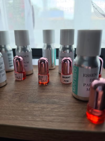 This image shows a collection of small perfume sample bottles arranged on a wooden surface. The bottles are a mix of clear glass and metallic silver, with some featuring pink metallic caps. Several bottles have white labels with black text, some of which are partially legible. One label clearly reads "MARSO F174". In the background, a sheer curtain is visible, suggesting an indoor setting near a window. The overall lighting is soft and diffused. There are no people visible in the image, and no actions are depicted. The environment appears to be a domestic or retail display setting. There is no information within the image that can determine the location in the city of Krasnochervonnyi, Russia.