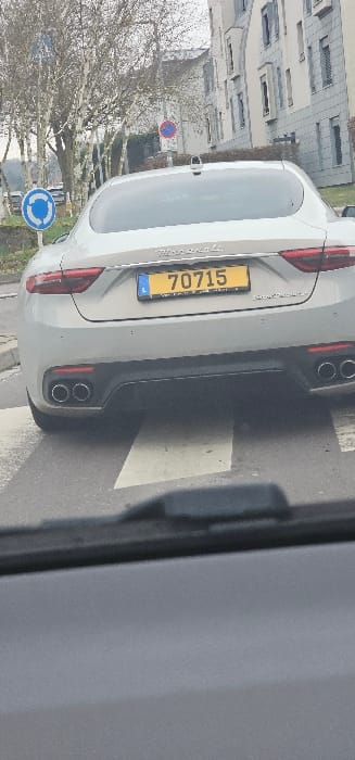 The image displays the rear view of a silver Maserati GranTurismo stopped on a pedestrian crossing in Dudelange, Luxembourg. The vehicle exhibits a yellow Luxembourgish license plate, number "L 70715", centered on its trunk lid, below which "Maserati" and "GranTurismo" badging are visible. The lower rear bumper features a dark diffuser section and four chrome-tipped exhaust outlets. The viewpoint is from inside another vehicle, indicated by the dark dashboard and a windshield wiper visible at the bottom of the frame. The immediate environment includes white zebra stripes of the pedestrian crossing on dark asphalt. In the background, to the left, are several leafless deciduous trees and a blue circular road sign indicating a roundabout. To the right, multi-story modern residential buildings with light-colored facades and windows are present, alongside a circular red and blue 'no stopping' road sign. The scene is under overcast lighting conditions. No people are visible.