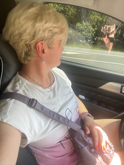 In Samylenka, Russia, a woman with blonde hair is sitting in the passenger seat of a car, looking out the window. She is wearing a white t-shirt with a gradient pink design and a purple purse is slung across her body. A necklace with a circular pendant and a butterfly earring are visible. Her hand, with bright orange nail polish, rests on the purse. Outside the window, trees and vegetation line a road with white lane markings. A reflection on the window shows a woman with blonde hair and bright orange nails, and a purple purse, suggesting it might be a reflection of the woman in the car, or another passenger.
