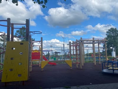 The image depicts an outdoor playground on a cloudy day. In the foreground, a yellow climbing wall is visible, angled to the left. To the right of the climbing wall are various playground structures, including monkey bars, a slide with red and yellow accents, and a small blue and yellow merry-go-round. A metal fence runs across the background, separating the playground from a grassy area with trees and some distant structures, including a statue. The ground of the playground is covered with dark, impact-absorbing material. The sky is predominantly blue with large, white cumulus clouds. This scene is located in the village of Nadezhdino, Russia.