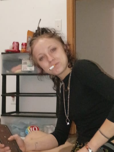 A woman is holding a cigarette in her mouth. She is wearing a black long-sleeved shirt and a chain necklace. She is indoors and there are storage containers, some with visible labels, behind her. The location is Clarksville, United States of America.