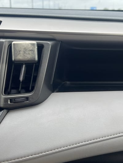The image shows a close-up of a car's interior. Visible are the air vent, a car fragrance diffuser attached to the vent, the car door trim, and a portion of the side window. The fragrance diffuser is rectangular and appears to be made of metal. The door trim is upholstered with white material and features stitching. The location is not discernible from the image.