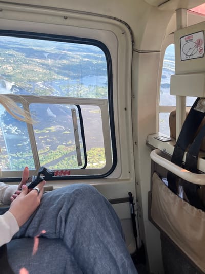 The image depicts the interior of a helicopter during a flight over a landscape. The helicopter's window shows a view of green terrain with a river. The interior of the helicopter reveals a passenger, identifiable by their grey pants, beige shirt, hands, and a section of blonde hair. One hand holds a lever attached to a smaller openable window. A warning sign is visible in the upper left corner of the window frame. The location can be inferred from the landscape to be in the vicinity of Victoria Falls Municipality, Zimbabwe.