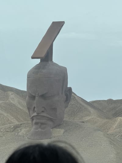 The image displays a massive, light brown stone sculpture of a human head, depicted in profile and facing slightly upwards and to the left. The head appears to be male, characterized by a dignified or contemplative expression with discernible facial features including a nose, mouth, and eye. Integrated into the back and top of the head is a large, rectangular, reddish-brown structure, which resembles an open book or a supporting plinth, extending upwards and backward.

The sculpture is set outdoors against a rugged, textured background of natural rock or carved stone, which shares a similar muted brown and gray color palette with the sculpture itself, suggesting a cohesive artistic integration into the landscape. Above the sculpture and the rocky terrain, a pale, overcast sky dominates the upper portion of the frame, indicating a daytime setting, possibly under hazy or cloudy weather conditions, as there are no distinct shadows.

The scene is static, capturing a monument or art installation. The grandeur and scale of the sculpture, combined with the austere landscape, evoke a sense of monumentality. This type of large-scale land art is consistent with the provided location context of Guazhou County, China. No activity is taking place, and there is no visible text. A dark, blurry obstruction is present in the lower right corner of the original image, partially obscuring the view.