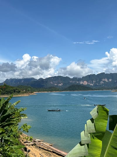 The image depicts a serene natural landscape in Ban Ta Khun, Thailand. A vast, tranquil turquoise lake stretches across the frame, bordered by lush green mountains and hills. The sky is a brilliant blue, dotted with fluffy white clouds.
In the foreground, large, vibrant green banana leaves frame the scene, adding a tropical feel. Along the shoreline, a mixture of sandy and rocky terrain is visible, with some fallen logs and debris suggesting a natural, untouched environment.
Several small boats navigate the lake. One dark, long-tailed boat is closer to the shore, while other smaller boats are seen further out, creating white wakes on the water's surface. The mountains in the background are covered in dense vegetation, with some lighter-colored rock faces visible, and a few scattered structures can be seen nestled among the trees on the hillsides. The overall atmosphere is peaceful and idyllic, capturing the natural beauty of the region.