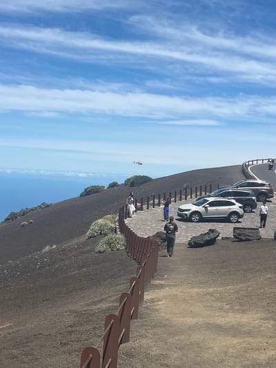 The image depicts a scenic overlook in La Orotava, Spain, likely at the Roques de Garcia or a similar volcanic landscape. In the foreground, a rustic metal railing follows a paved path that curves along a dark, volcanic slope. Several large, dark rocks are scattered on the ground.

Further along the path, people are visible. One individual, wearing dark clothing, walks away from the camera. Another person in a white dress is seen near a small group.

A line of parked cars, including a white SUV and a dark SUV, are situated on a paved area to the right. One white car, a compact SUV, is prominently featured. A man in a white shirt and black pants stands next to one of the cars.

The background reveals a vast expanse of blue ocean meeting a hazy horizon under a bright blue sky dotted with wispy white clouds. A small yellow helicopter is captured in mid-flight in the upper portion of the image, above the ocean. The landscape is characterized by its dark, earthy tones, contrasting with the vibrant blue of the sea and sky.