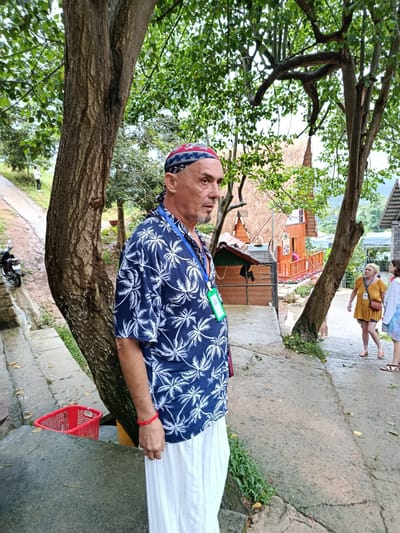 A man with a bandana and a blue short-sleeved shirt with a palm tree print stands on a wet, paved pathway. He is wearing white striped pants and a red bracelet on his left wrist. A lanyard with green and white tags is around his neck. To his left, a red basket is visible on a grey concrete ledge. In the background, there are trees, buildings with thatched roofs, and other people walking on the path. A motorcycle is parked to the left of the man. The scene appears to be in an outdoor tourist area in Định An, Vietnam.