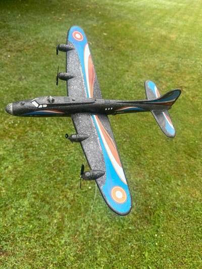 A black toy airplane is positioned outdoors on a patch of green grass. The airplane has four propellers, two on each wing. The wings and tail are decorated with blue and orange stripe patterns and circular insignia. The scene appears to be in Lapės, Lithuania, as indicated by the provided metadata. The grass is well-maintained and appears to be a lawn. The lighting suggests it is daytime, with no specific weather conditions discernible. There are no people or animals visible in the image.