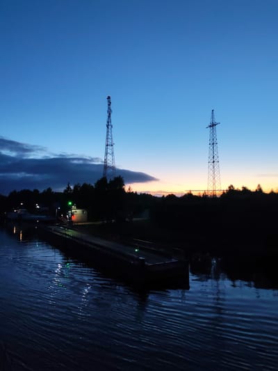 The image shows Annensky Most, Russia at twilight. The sky is a gradient of deep blue to a pale orange on the horizon. Two lattice towers stand silhouetted against the sky. A dark, elongated object, possibly a barge, is visible in the water with green lights reflected. A small building with green lights is situated near the barge. A bank of trees and dark clouds are positioned to the left. The water surface is rippled.