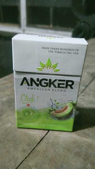 The image shows a pack of Angker cigarettes with "AMERICAN BLEND" written on it. The pack is mostly white with green accents, featuring a leaf logo and an image of a sliced melon with water splashes. Text on the pack indicates "MADE UNDER AUTHORITY OF UFA TOBACCO INC. USA" and "20 PREMIUM TOBACCOS". The cigarette pack is placed on a tiled surface, with a dark background that suggests an indoor setting. There are no people visible in the image. The location is not specified in the image itself, and therefore its presence in Wonosegoro, Indonesia cannot be confirmed.