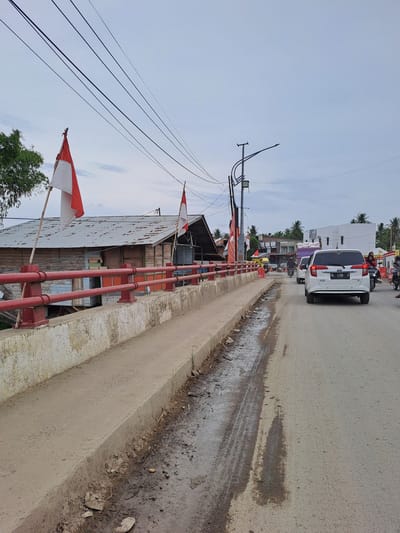 The image captures a street scene in Lhoknga, Indonesia. A paved road with tire tracks and patches of standing water runs diagonally from the lower right to the upper left. A white SUV with license plate "BL 1498 K" is in the foreground, driving away from the viewer. To the left of the road is a concrete embankment with a red metal railing. Behind the railing, a rustic wooden building with a corrugated metal roof is visible. Two Indonesian flags, red and white, are mounted on bamboo poles on either side of the building. Further down the street, traffic including motorcycles, cars, and a truck with a load of what appears to be sacks can be seen. The background is lined with palm trees and buildings, with a cloudy sky overhead.
