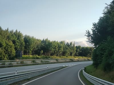 The image shows a highway in the city of Oldenburg in Holstein, Germany. The highway has two lanes in each direction, separated by a median strip. The lanes are paved with asphalt, and there are white lines marking the edges of the lanes. There are metal guardrails along the sides of the highway. On either side of the highway, there are trees and vegetation. The sky is clear and blue, with some clouds in the distance. There are no people or vehicles visible in the image. The scene appears to be peaceful and natural, with the highway cutting through the landscape.