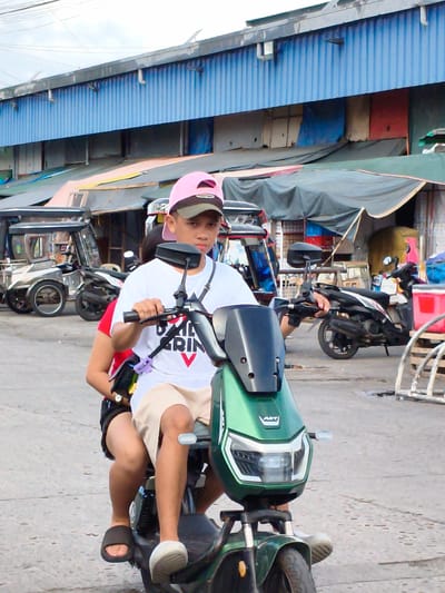 Two people are riding on a green electric scooter. The person in front, a young male wearing a pink baseball cap, a white t-shirt with a graphic that appears to say "AXIL GRIN V", and tan shorts, is driving. The person behind him, wearing a red shirt and black shorts, is seated. The scooter has a black windshield and a black storage compartment. The scene appears to be an outdoor market or street, with various stalls and structures visible in the background. These include a blue corrugated metal building with awnings and tarps, suggesting a commercial area. A tricycle is parked to the left, and other motorcycles are visible in the background. The lighting suggests it is daytime, with overcast skies. The overall atmosphere is casual and typical of a busy street scene in the Philippines. There is no clear indication of a specific event, but it appears to be a normal day of transit or activity within the community.
