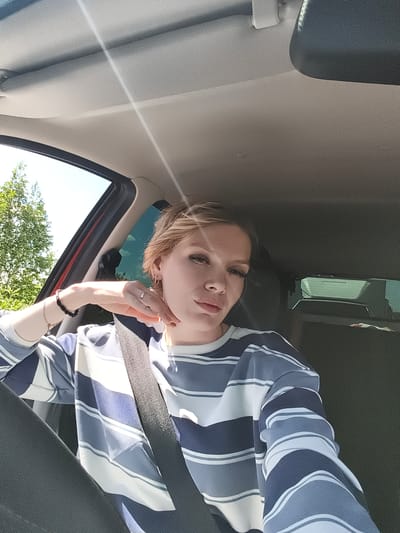 A young woman with blonde hair and makeup is taking a selfie in the driver's seat of a car, on a sunny day, with trees visible through the window. She is wearing a striped sweater and a seatbelt. Her left hand is resting on her face and her right arm is resting on her lap. The interior of the car, including the ceiling and dashboard, is visible. The location is the city of Yagul, Russia.