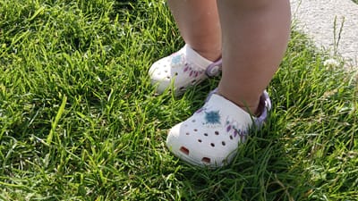 In Sankt Pölten, Austria, a close-up shot shows a child's legs and feet wearing white Crocs adorned with purple and teal diamond-shaped patterns and a teal snowflake-like embellishment. The child is standing on vibrant green grass, which is the dominant element of the environment. A hint of a light-colored, textured surface, possibly a paved area or stone, is visible in the upper right corner, along with a few thin blades of grass reaching upwards. The lighting suggests it is daytime, with sunlight illuminating the scene and casting subtle shadows.