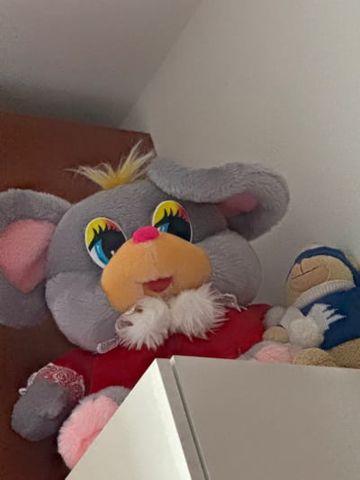 The image shows two stuffed animals on a shelf. The larger stuffed animal is gray with pink ears, a large orange snout, and big colorful eyes. It is wearing a red garment with white fur trim. A smaller tan stuffed animal wearing a blue and white patterned scarf is partially visible to the right of the larger stuffed animal. The background consists of a brown wall and a white wall, with a corner where they meet. The shelf appears to be made of a light-colored material, possibly wood or laminate. The overall setting suggests an indoor environment, potentially a child's bedroom or playroom. There is no indication of a specific location in Borgosatollo, Italy, in this image.