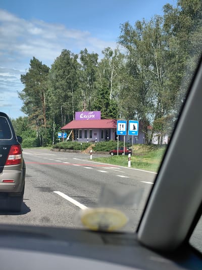 The image captures a roadside scene on a partly cloudy day in Бараўскі сельскі Савет, Belarus, viewed from inside a vehicle. In the foreground, the rear of a dark gray or brown SUV, possibly a Kia Sorento (indicated by visible "EX" badging), is partially visible on the left. A prominent red horizontal line is painted across the asphalt road, functioning as a stop line or demarcation for an intersection or crossing.
To the right of the road, a single-story building with light purple exterior walls and a red tiled roof stands behind a small landscaped area with trimmed green bushes. A large purple sign with the word "Сябры" (Syabry, meaning "Friends" in Belarusian) in white Cyrillic script is mounted on the roofline of the building. In front of the building, two blue square informational road signs are erected on poles: the left sign displays a fork and knife pictogram, indicating a restaurant or food service, and the right sign displays a pine tree pictogram, signifying a recreational or forest area, both accompanied by right-pointing arrows. A small, dark red sedan is partially visible, parked beyond the delineator post and partially obscured by the building and grass. A white and red striped delineator post is positioned on the road's shoulder.
The background is dominated by a dense line of trees, including both light-barked deciduous trees (likely birches) and darker conifers (likely pines), extending across the horizon. The sky is blue with scattered white cumulus clouds.