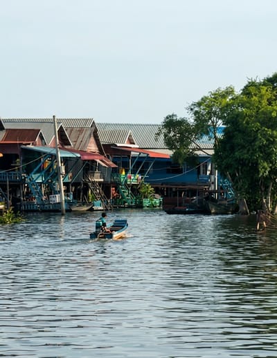 A young person in a blue shirt and dark shorts is operating a small, motorized blue and orange boat that is moving from left to right across calm, rippling water. The boat is leaving a wake behind it. In the background, a stilted village with buildings featuring red and corrugated metal roofs lines the waterfront. One building is painted bright blue. Green vegetation and trees are visible behind the structures, and the sky above is clear and pale blue. The scene is set in the stilted village of Kampong Khleang, located in the Soutr Nikom district of Siem Reap Province, Cambodia.