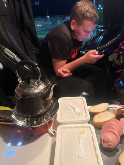The image depicts a young man in a dark t-shirt sitting in what appears to be the cabin of a vehicle, possibly a truck, at night. He is looking down at his phone, which he is holding in his hands. The cabin is dimly lit, with some lights visible from the exterior, suggesting he is stopped somewhere. In the foreground, there are several objects laid out on a surface: a stainless steel kettle on a red portable stove, two white plastic containers with forks, slices of bread, and a pink, sliced sausage. A digital display shows "290." The overall scene suggests a break during a journey, with the man using his phone while food is being prepared or has been laid out. The location is in the city of Lukhovitsy, Russia.