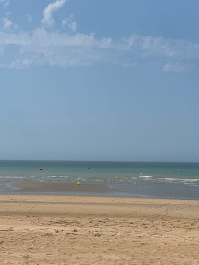 This is a vertical, eye-level, medium shot of a beach scene. The image is divided into three distinct horizontal sections: the sky, the sea, and the sand.

The top section is a clear, pale blue sky with a few wispy white clouds on the left side, suggesting it's daytime and the weather is fair.

The middle section depicts the sea, appearing calm and shallow near the shore. The water is a muted turquoise color. Several yellow buoys are visible floating on the surface of the water, spaced out in a line. Further out in the sea, two small red objects, possibly kites or buoys, are also visible. The waves are gentle, creating a subtle white line where they meet the sand.

The bottom section is a sandy beach, exhibiting a light brown, granular texture. The sand appears dry and shows faint footprints, indicating recent human activity. The beach stretches horizontally across the frame, meeting the water's edge.

The overall mood is peaceful and serene, capturing a tranquil day at the beach. The scene is likely set in Koksijde, Belgium, as indicated by the context. There is no visible text in the image.