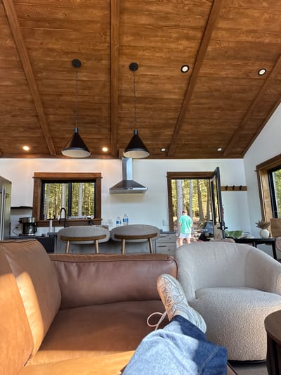 The image displays the interior of a modern living space or cabin located in Otego, United States, looking towards a kitchen area and an outdoor environment. In the immediate foreground, the lower legs and feet of a person are visible, resting on a brown leather sofa. The person is wearing blue jeans and white sneakers. To the right of the sofa, a cream-colored, textured armchair is partially visible.
The ceiling features warm-toned wooden planks with exposed beams, illuminated by several recessed spotlights and two black conical pendant lights suspended by chains. The walls are painted white.
The mid-ground reveals a kitchen counter area with a dark sink, a black faucet, and a coffee maker on the left. A stainless steel refrigerator is positioned on the far left. Two modern bar stools with light gray upholstered seats and wooden backs are placed at the counter. A stainless steel range hood is mounted on the wall above the counter, where two bottled water bottles are also present.
Through a large window above the kitchen sink and another window on the far right, a dense green forest is visible. An open doorway on the right side leads outdoors, through which a person, seen from behind, is walking away from the camera. This individual is wearing a light teal or mint green short-sleeved shirt and white shorts, heading into the sunlit wooded area. A small vase containing dried floral elements sits on a counter surface on the far right, near the doorway. A hook rack is mounted on the wall adjacent to the open door.