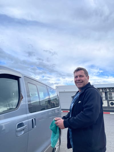 A man is polishing a silver van in an outdoor urban setting. The sky is cloudy, with white and blue hues. The man is wearing a dark jacket. Multiple air conditioning units are visible in the background. The location is identified as Бавыкино, Russia.